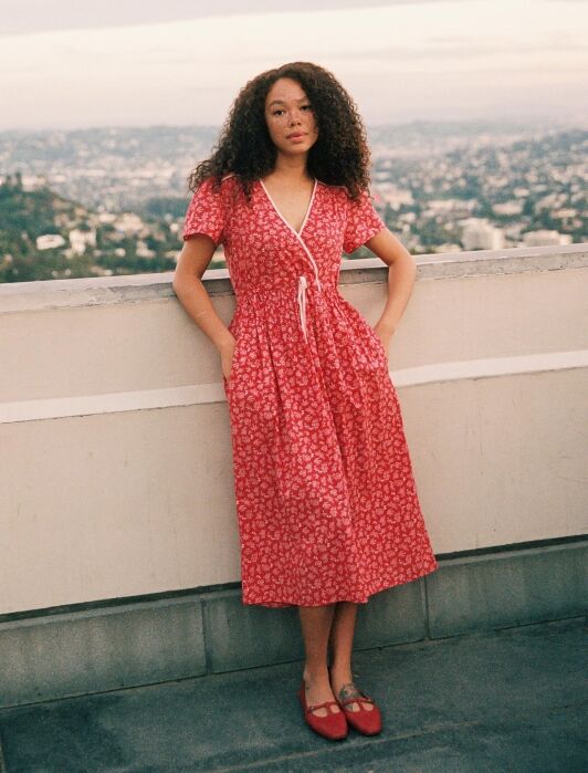 A woman in a red patterned dress stands by a rooftop ledge with a cityscape in the background.