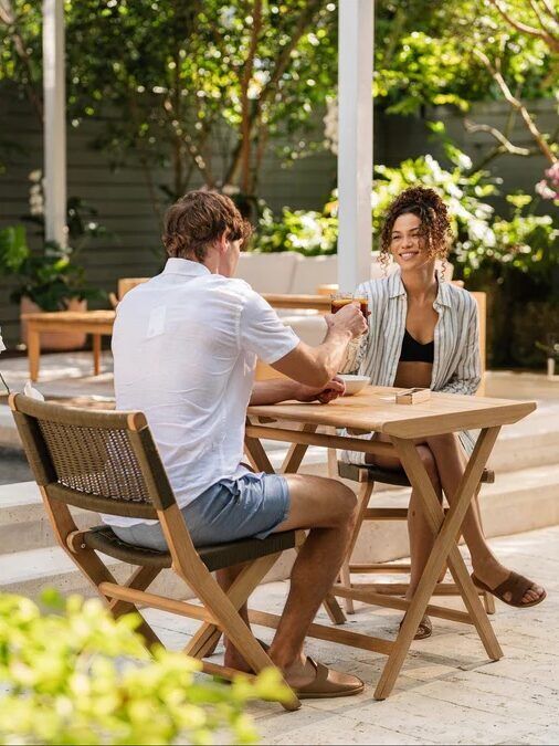 Two people sit at a wooden table outdoors near a pool, smiling and holding hands, surrounded by green plants and patio furniture.