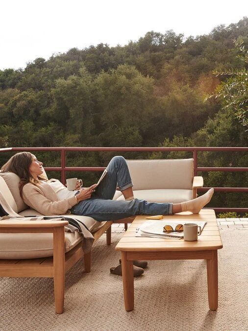 A person lounges on a wooden sofa on a balcony, reading a book, with coffee cups and plants nearby; surrounded by chairs and overlooking a green, wooded landscape.
