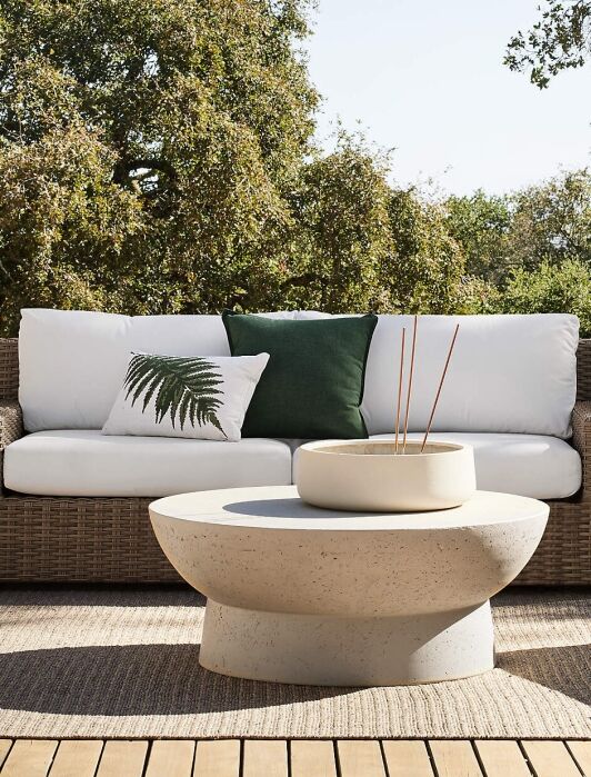 A wicker outdoor sofa with white cushions and two green pillows sits on a wooden deck behind a round stone coffee table, surrounded by trees.