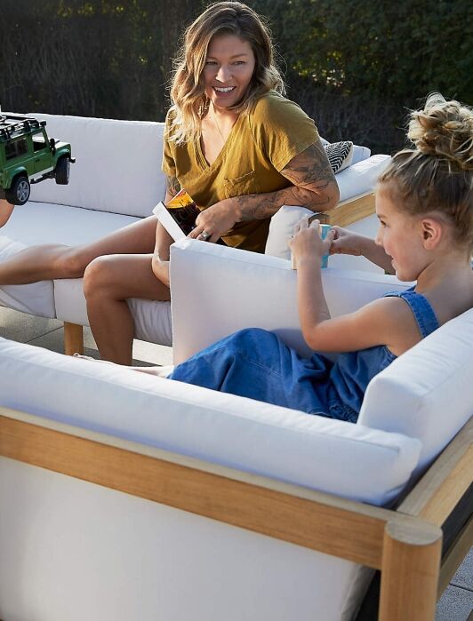 A woman sits on an outdoor sofa set, smiling and interacting with two young children playing with toys on a patio.