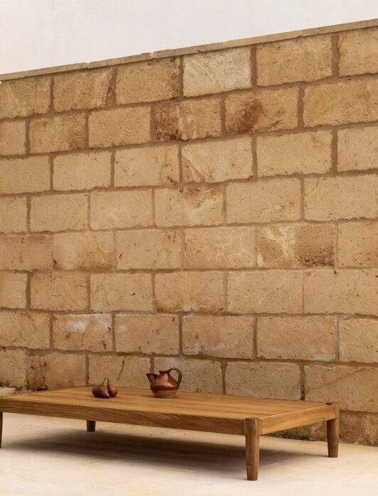 A low wooden table with two small ceramic pitchers sits in front of a stone wall next to a glass door and some rocks outside.