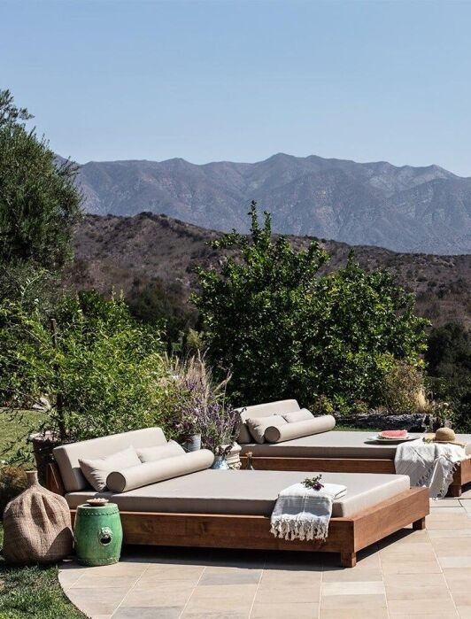 Two outdoor lounge beds with cushions and towels are positioned beside a pool, with mountains and greenery visible in the background under a clear sky.