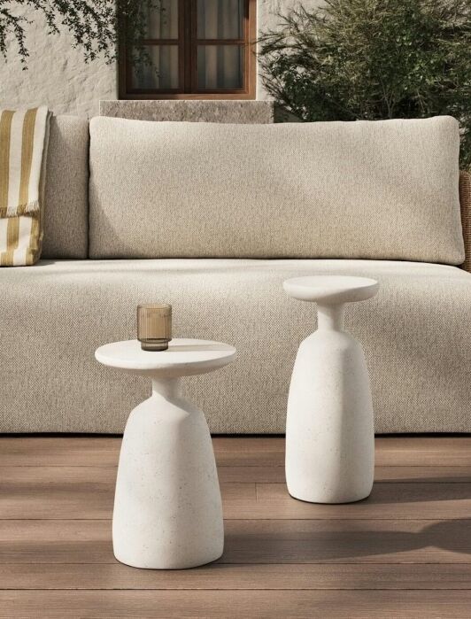 A beige outdoor sofa with a striped cushion behind two white modern side tables, one holding a glass, on a wooden deck with greenery and a window in the background.