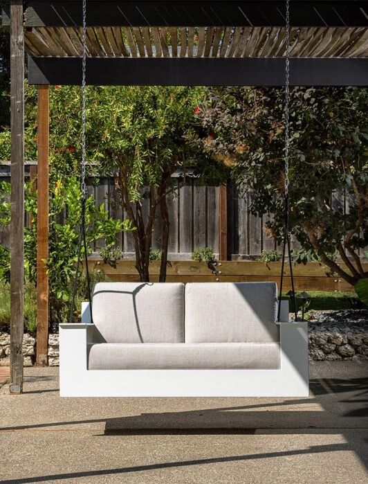A modern white porch swing with beige cushions hangs from a pergola in a landscaped backyard with trees, plants, and a wooden fence.