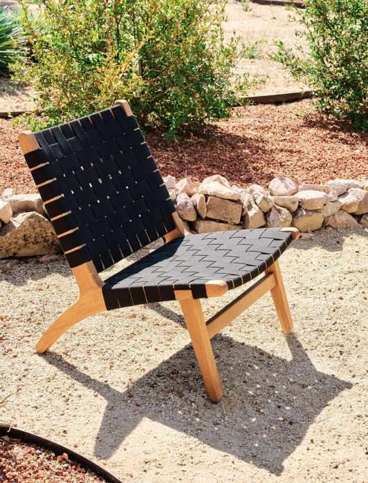 A wooden chair with a woven black seat and backrest is placed outdoors on a sandy surface, surrounded by rocks and plants.
