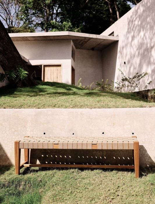 A wooden bench sits on grass in front of a beige concrete wall, with a modern house and trees visible in the background.