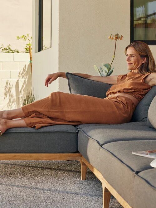 A woman sits relaxed on a gray outdoor sectional sofa, leaning back with her legs stretched out on the seat, in a bright patio area with plants and neutral walls.