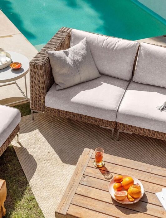 Outdoor patio set with wicker sofa and chair, a wooden coffee table with oranges and drinks, a white side table with candles, and a swimming pool in the background.