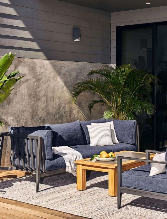 Modern outdoor patio with a wooden coffee table, gray cushioned seating, decorative pillows, potted plants, and a rug on a wooden deck next to a glass door.