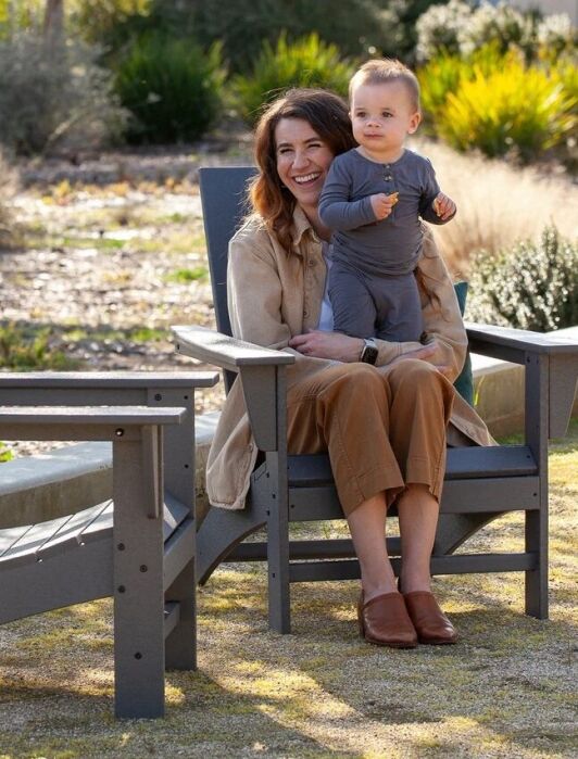 A woman sits on an outdoor chair holding a baby on her lap, with an empty chair beside them in a garden setting.