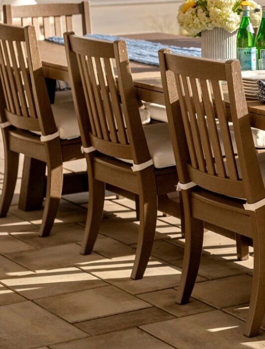 Four wooden chairs with seat cushions are arranged along one side of an outdoor dining table set with bottled drinks, a vase of flowers, and folded napkins on a tiled patio.
