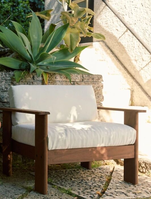 A wooden outdoor bench with white cushions sits beside a large terracotta plant pot and green leafy plants, against a stone wall in a sunlit garden.