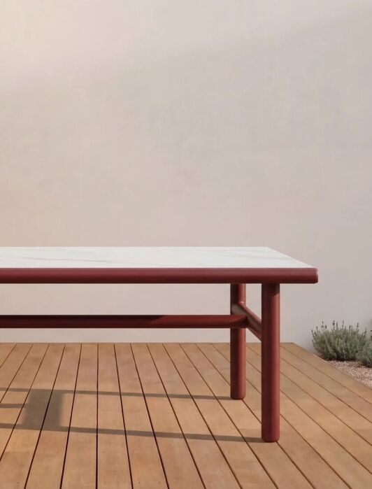 A rectangular table with a white top and red legs stands on a wooden deck, surrounded by small plants and next to a light-colored wall.