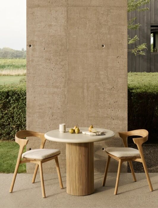 A round table with two wooden chairs is set with plates, cups, and pears on a patio beside a concrete wall, with greenery and a modern dark building in the background.