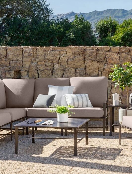 Outdoor patio set with beige cushions, black metal frames, and a coffee table, placed in front of a stone wall and surrounded by greenery on a sunny day.