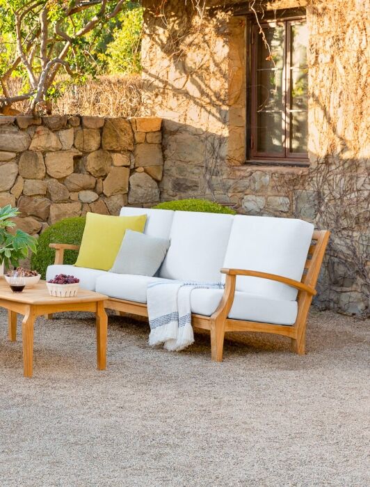 Outdoor patio area with wooden furniture featuring white cushions, a stone fireplace, a coffee table with decor, and various plants in a sunlit garden setting.