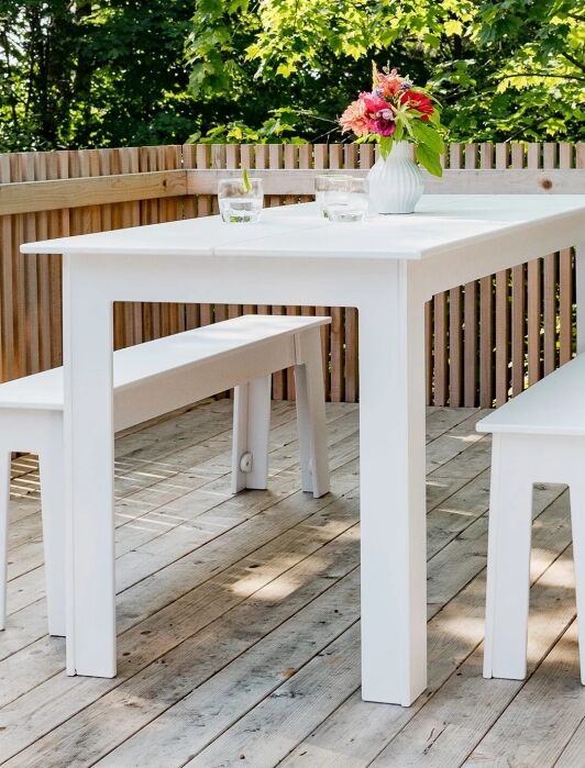A white outdoor table with two matching benches sits on a wooden deck, decorated with a vase of flowers and two glasses of water, surrounded by a wooden privacy fence and greenery.