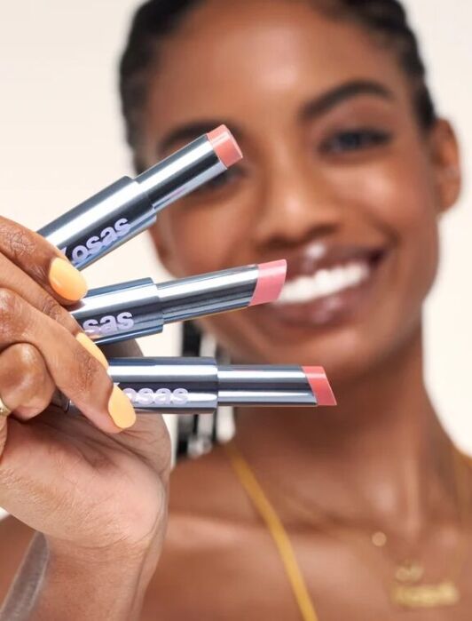 Person holding three tubes of lipstick against a blurred background. The lipsticks have different shades of pink. The person is wearing a yellow top and has braided hair.