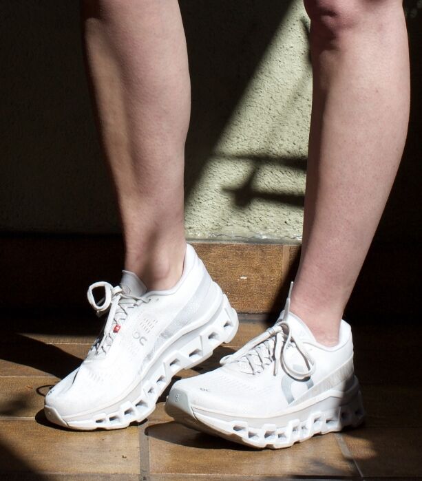 A person wearing white sustainable sneakers with uniquely shaped soles stands on a tiled floor in sunlight.