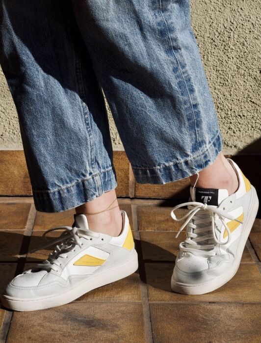 Person wearing loose blue jeans and white sustainable sneakers with yellow accents, standing on a brown tiled floor near a textured beige wall.