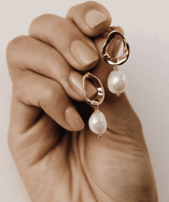 A hand with manicured nails holds a pair of gold earrings featuring irregular open shapes and dangling white pearls.
