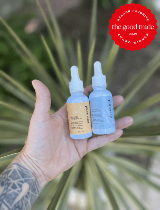 A tattooed hand holds two dropper bottles of Cocokind skincare serum outdoors, with a "The Good Trade 2026 Award Winner" badge in the top right corner.