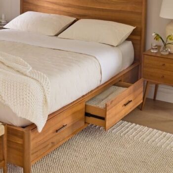 A wooden bed with white bedding features built-in storage drawers; one drawer is open and contains a basket. A knitted blanket and nightstand with a lamp are beside the bed.