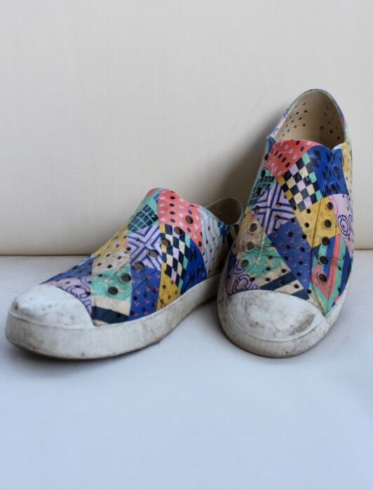A pair of sustainable sneakers in slip-on canvas style, featuring a colorful geometric patchwork pattern and visible signs of wear on the soles and toes.