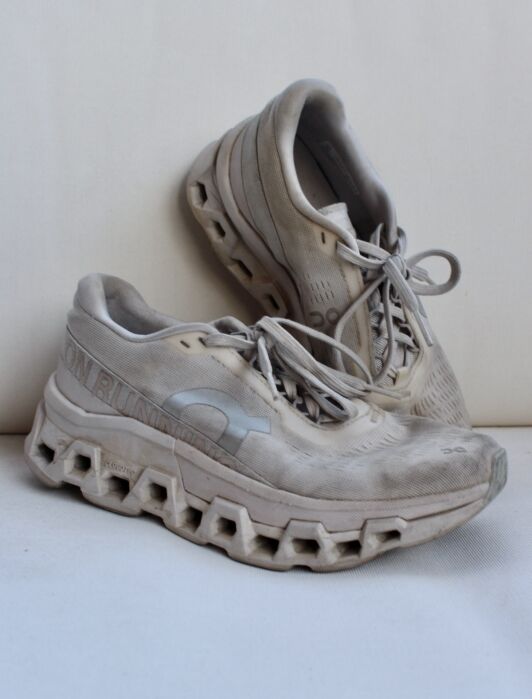 A pair of worn white sustainable sneakers with chunky, cut-out soles are stacked against a light-colored background.