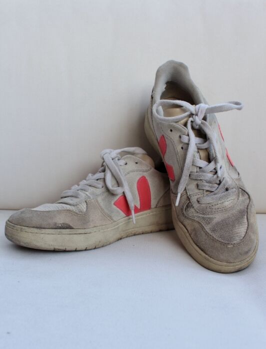 A pair of worn sustainable beige sneakers with red side logos and white laces, positioned against a plain off-white background.