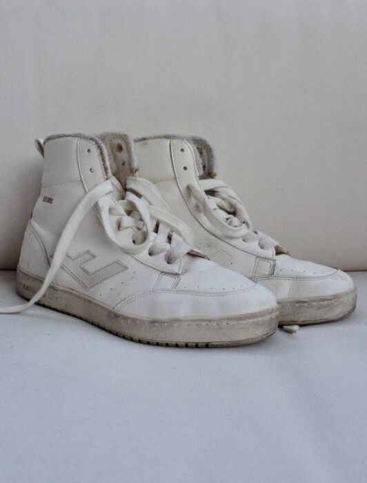 A pair of worn white high-top sustainable sneakers with visible scuff marks and one untied lace, placed on a light surface against a neutral background.
