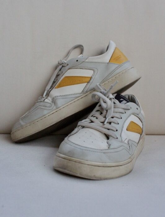 A pair of worn sustainable sneakers with white and yellow accents displayed against a plain light-colored background.