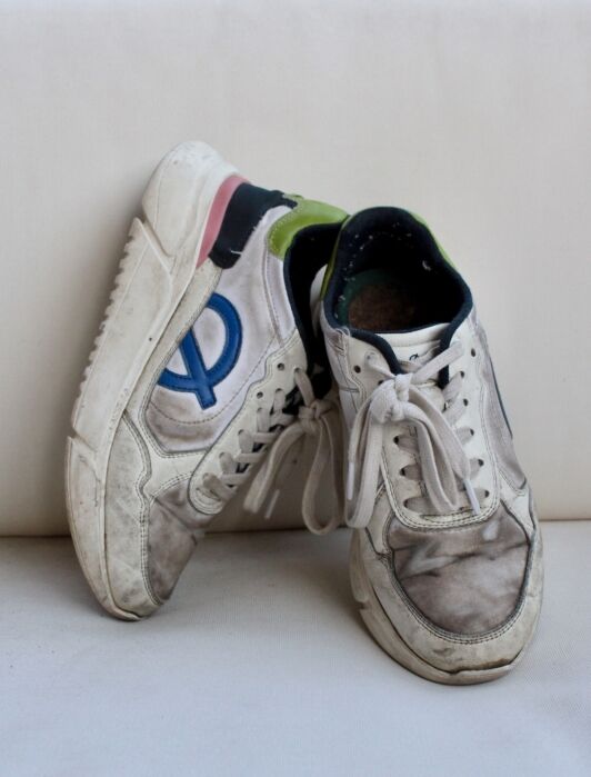 A pair of worn white sustainable sneakers with visible dirt, blue and green accents, and untied laces, resting against a white background.