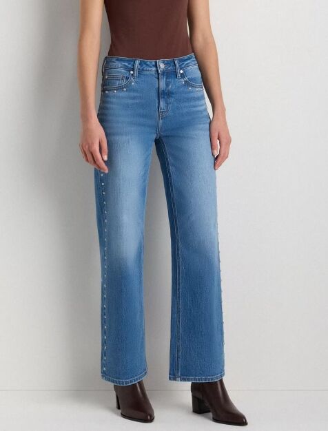 A person wearing light blue wide-leg jeans with rivet details on the side seams, a brown top, and brown ankle boots, standing against a plain light wall.