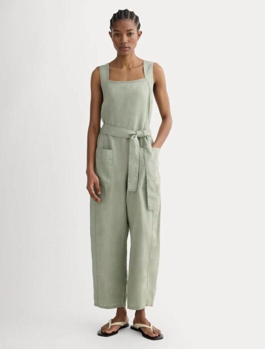 Person standing in a light green sleeveless jumpsuit with a belt and large front pockets, wearing black sandals against a plain background.