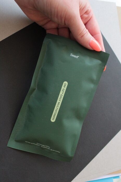 A hand holding a dark green Seed brand packet with text and a tear notch, positioned over layered sheets of paper.