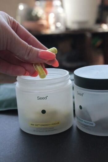 A hand holds two yellow-green capsules above an open Seed supplement container labeled "AM-02™ Energy + Focus." Another labeled container is nearby.