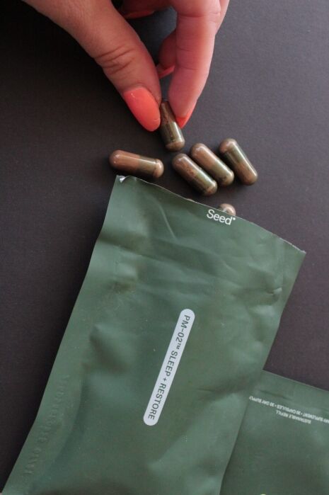 A hand with pink nails picks up a brown capsule near a green Seed-branded packet labeled "SLEEP RESTORE," with several capsules lying on the surface.