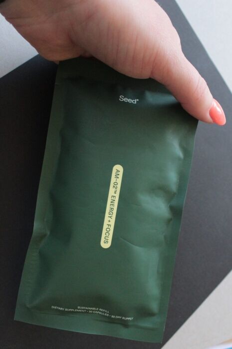A hand holds a dark green resealable pouch labeled “Seed AM+PM Energy + Focus,” described as a 30-day dietary supplement supply.