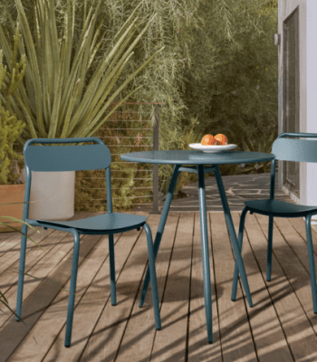 A small round table with two blue chairs on a wooden deck, with a plate of oranges on the table and plants in the background.
