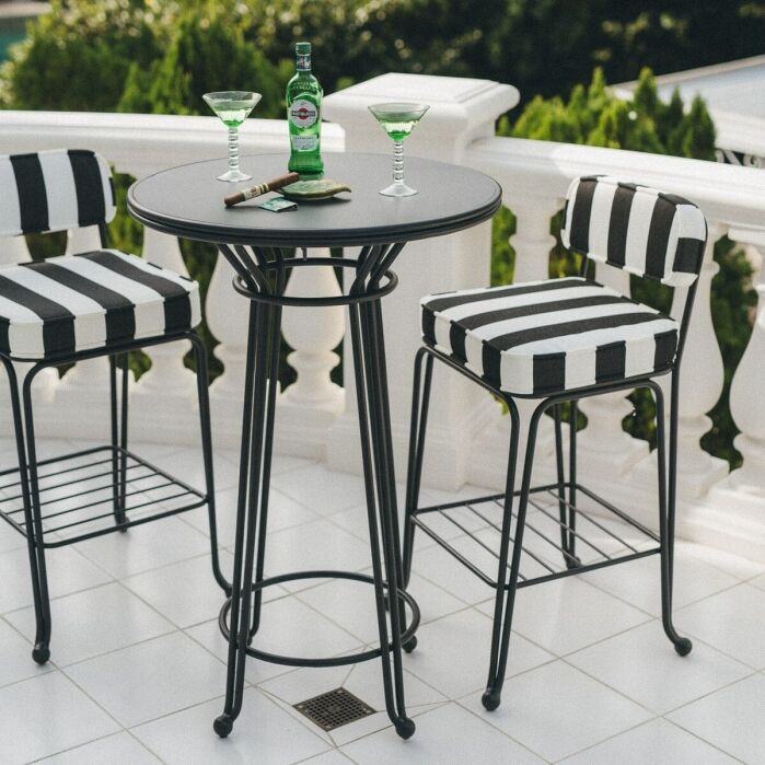 A black round table with two striped black-and-white chairs on a white tiled patio, featuring a green bottle, two cocktails, and sunglasses on the table.