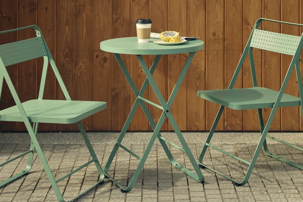 Two green metal chairs and a round table on a stone patio, with a coffee cup, a halved passion fruit, and a spoon on the tabletop, set against a wooden wall.