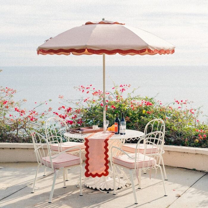A white metal patio table with four chairs sits under a pink umbrella, set with drinks and a patterned runner, overlooking the ocean with pink flowers in the background.
