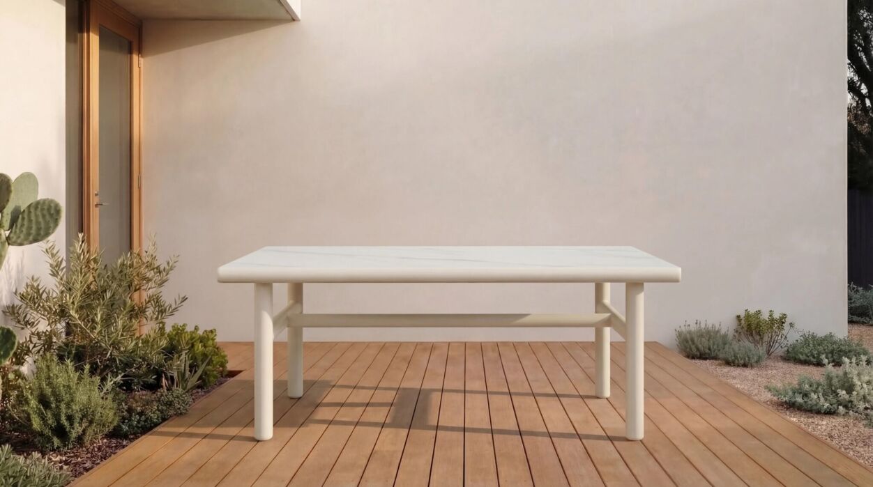 A white rectangular table stands on a wooden deck outside a modern house with light-colored walls, surrounded by plants and shrubs.