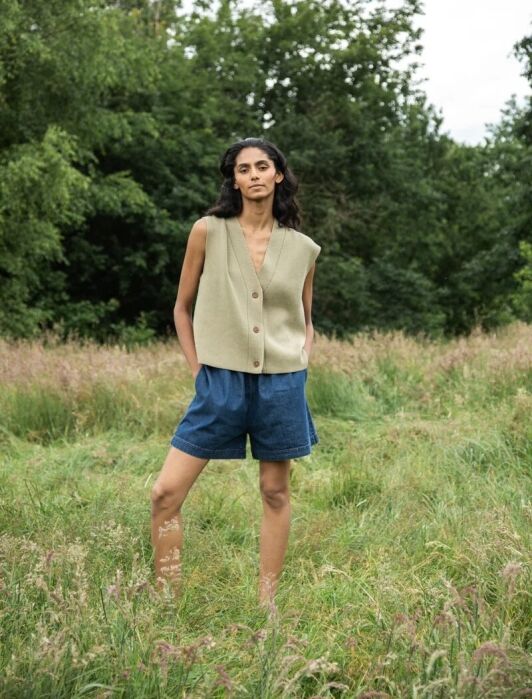 Person standing outdoors on grass, wearing a sleeveless buttoned vest and denim shorts, with green trees in the background.