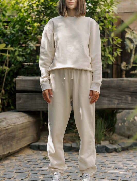 Person standing outdoors wearing a beige sweatshirt and sweatpants with white sneakers, set against a background of greenery and cobblestone pavement.