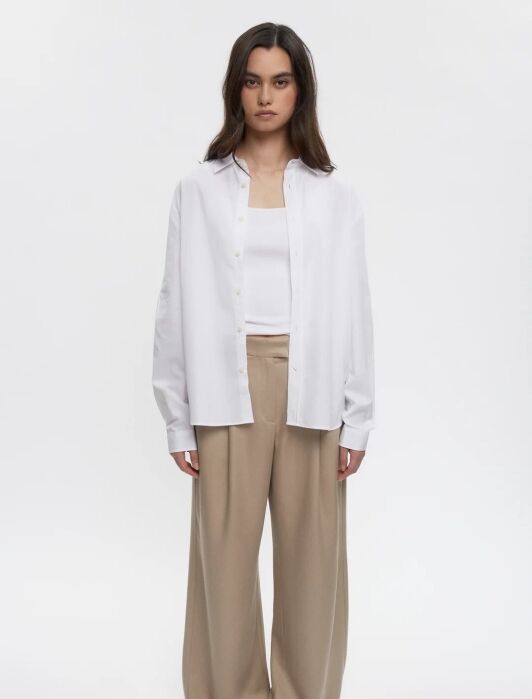 Woman standing, wearing a white shirt over a white top, beige wide-leg pants, and black sandals.