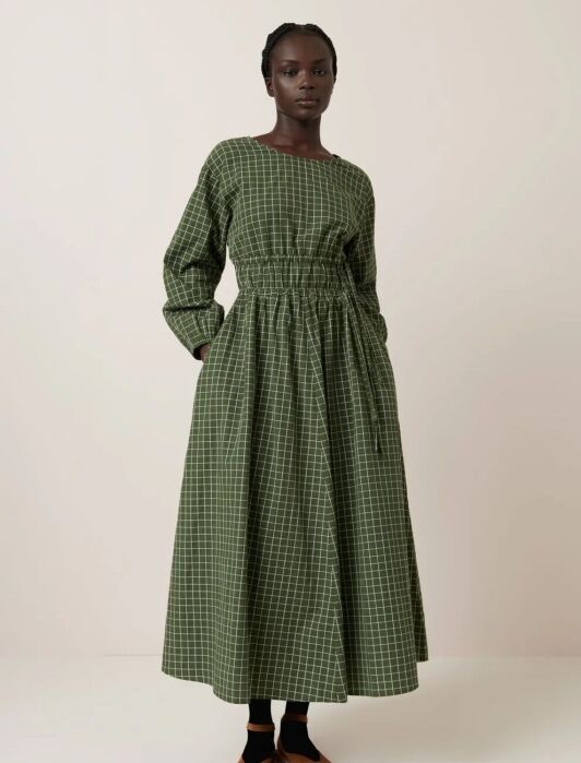 Person wearing a long-sleeved, green checkered dress with a cinched waist, black socks, and brown shoes, standing against a plain background.