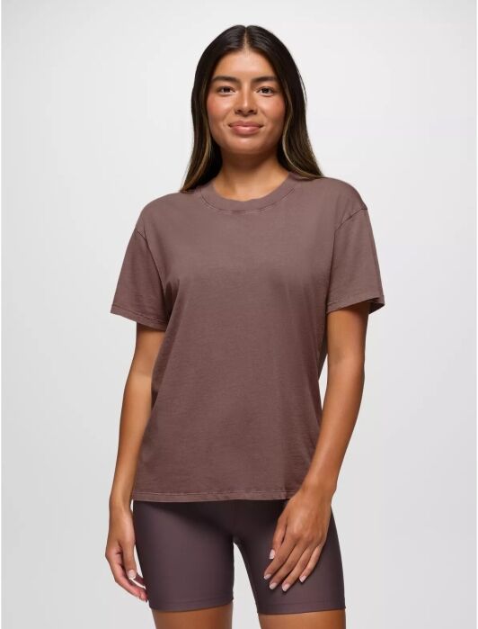 A person wearing a brown t-shirt and shorts stands against a neutral background, smiling with hands at their sides.